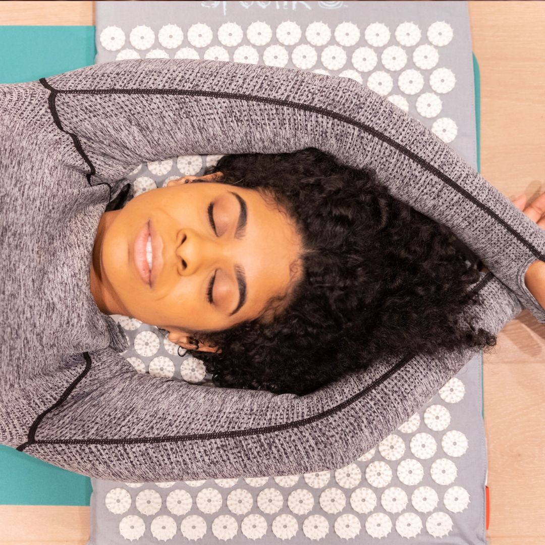 Woman with head laying on acupressure mat with arms stretched over head.