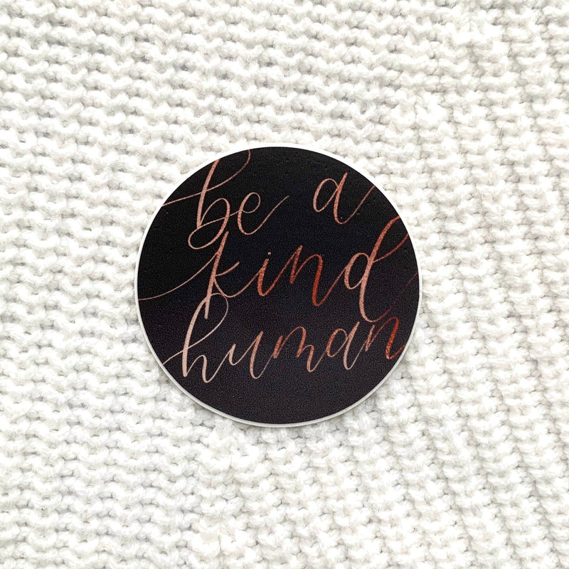 Round black sticker with "be a kind human" written in peach