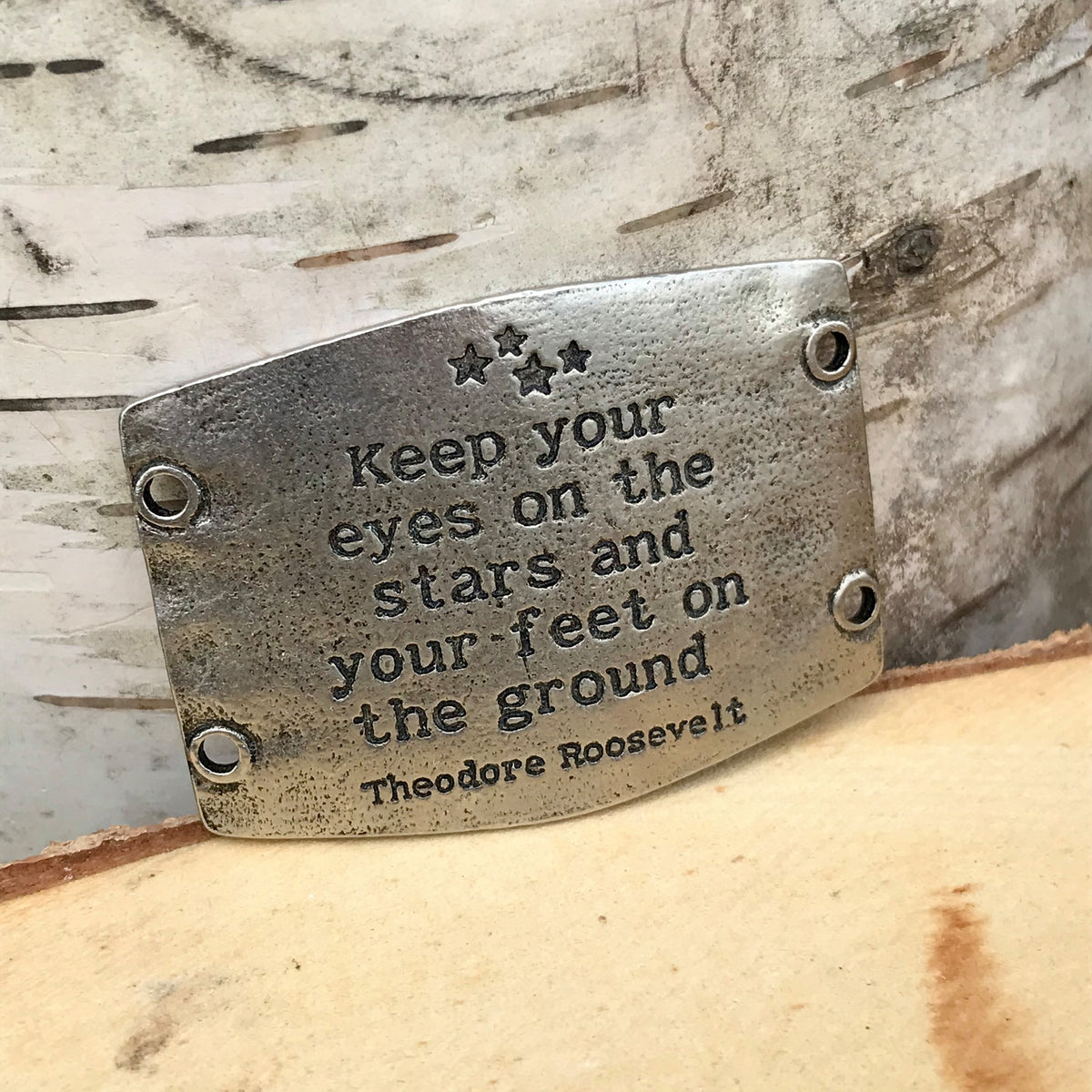 Antique silver finish Lenny & Eva bracelet sentiment that reads, "Keep your eyes on the stars and your feet on the ground." Quote by Theodore Roosevelt. Design of stars above the quote.