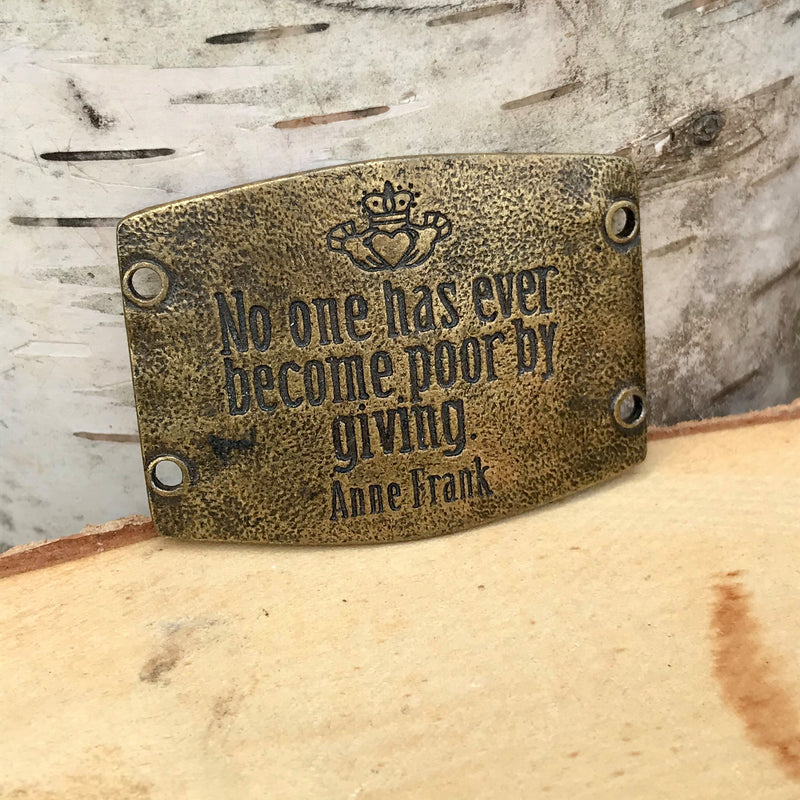 Antique brass finish Lenny & Eva bracelet sentiment that reads, "No one has ever become poor by giving." Quote by Anne Frank. Design above quote is two hands holding a heart and crown.