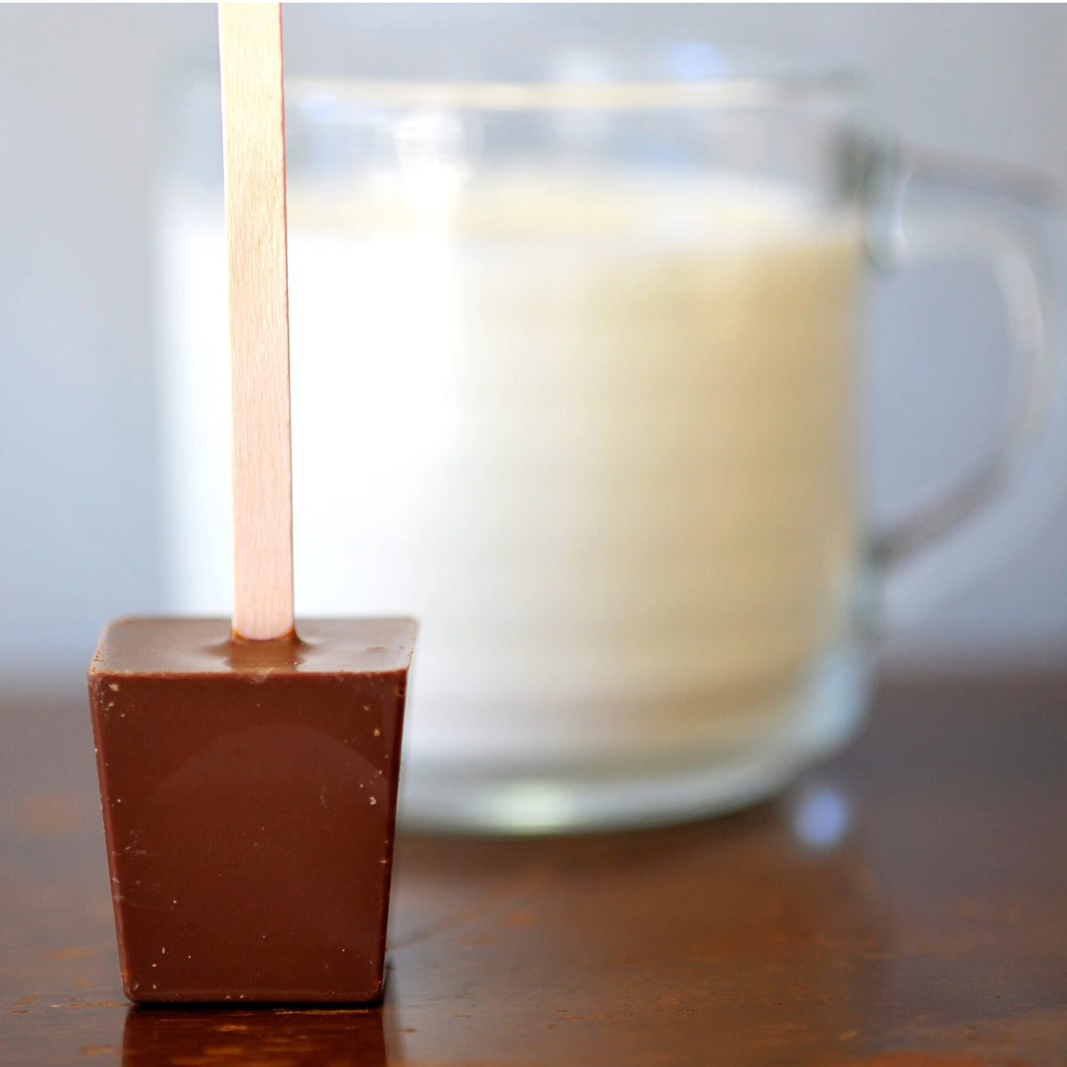 A cube of milk chocolate on a stick sitting in front of a mug of milk