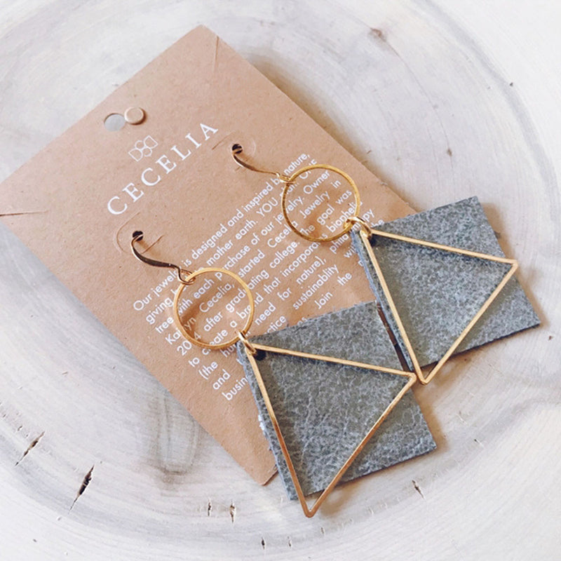Earrings with grey leather diamond shaped pendants hanging from gold circles and overlapped by gold triangles.