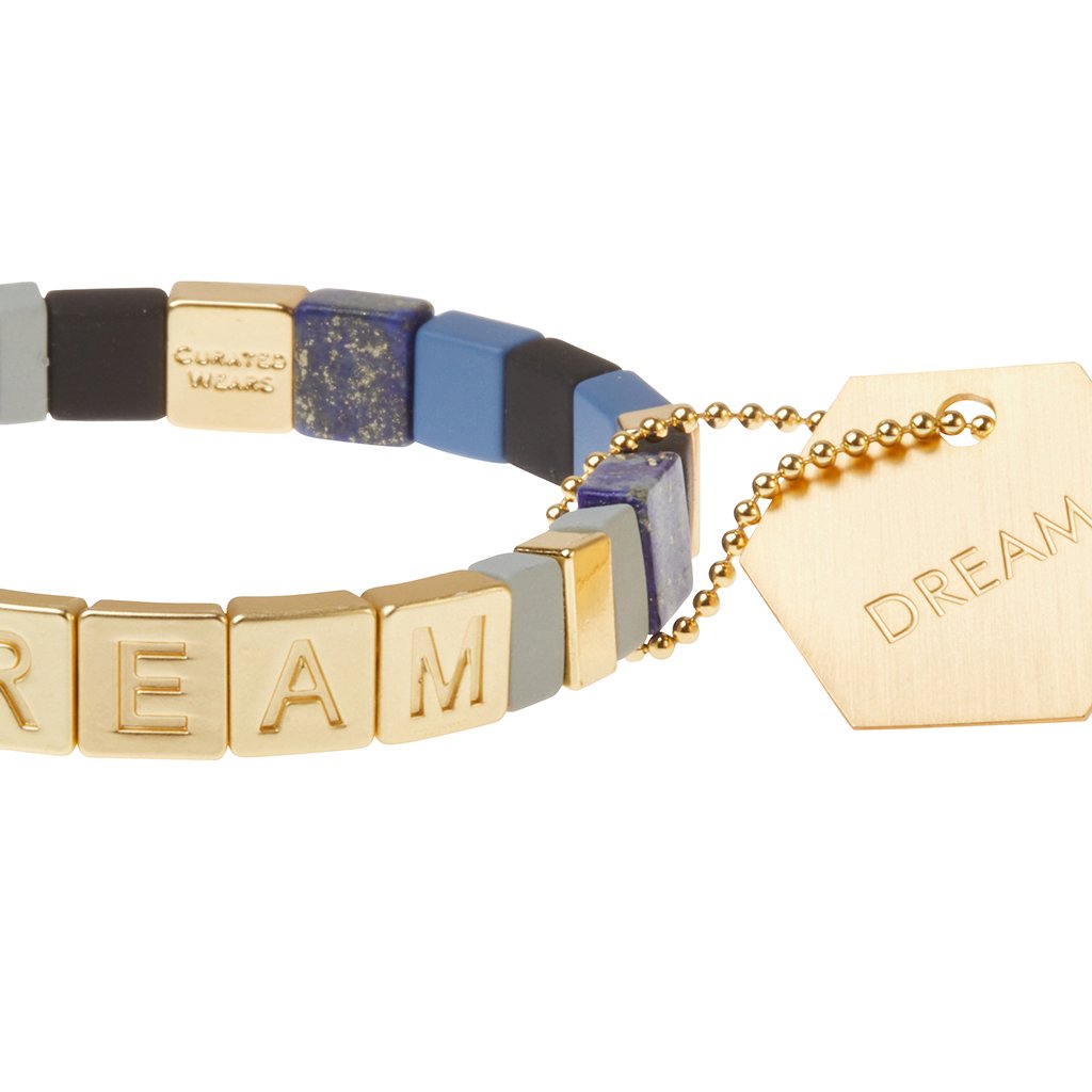 Close-up of blue, grey, black and gold beads on DREAM bracelet. Gold hexagonal DREAM stamped intention tag is attached.