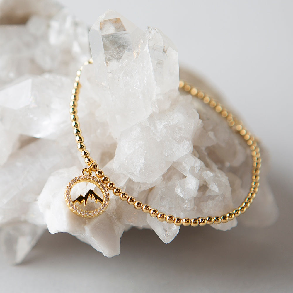 Lenny & Eva Keep Climbing bracelet. A bracelet with tiny gold beads for the band and a gold charm that's a circle of shiny faux diamonds with a mountain range silhouette in the middle.