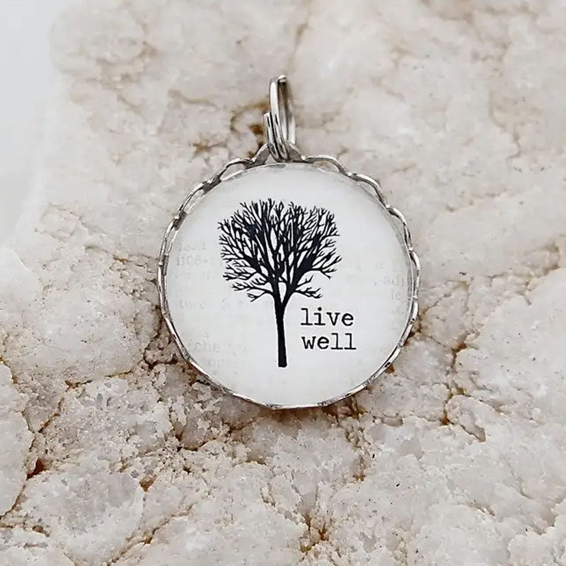 Round necklace pendant with a white background, silver metal edge, and image of a tree with the words "live well"