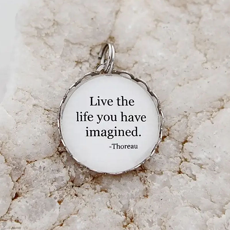 Round necklace pendant with a white background and silver metal edge that reads, "Live the life you have imagined. -Thoreau"