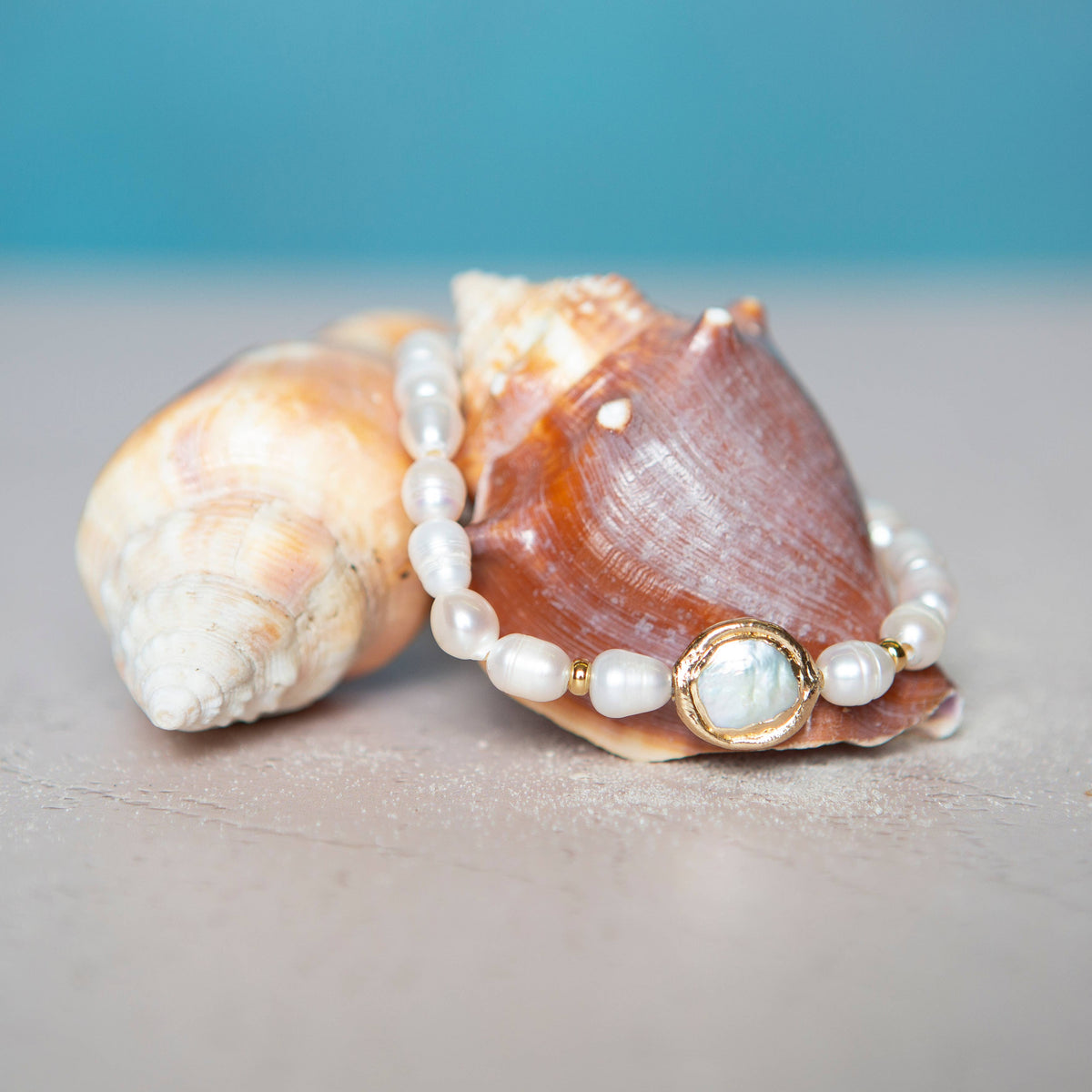 Lenny & Eva No Grit, No Pearl bracelet. A bracelet with irregular shaped freshwater pearl beads for the band and a circular focal pearl inside a gold colored circle. Bracelet is draped over a seashell