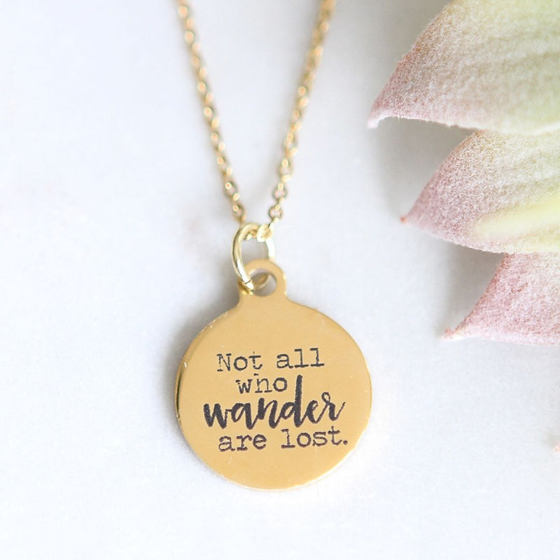 Round gold pendant engraved with "Not all who wander are lost" hangs from a gold necklace chain