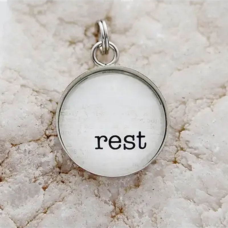 Round necklace pendant with white background and silver metal edge. Pendant reads "Rest".