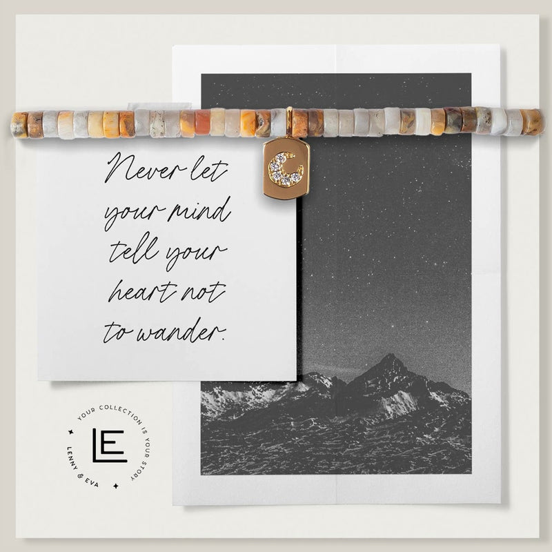 Bracelet with grey, brown, and rust colored Mexican Agate beads and a gold moon charm. On a card with a mountain background and text "Never let your mind tell your heart not to wander."