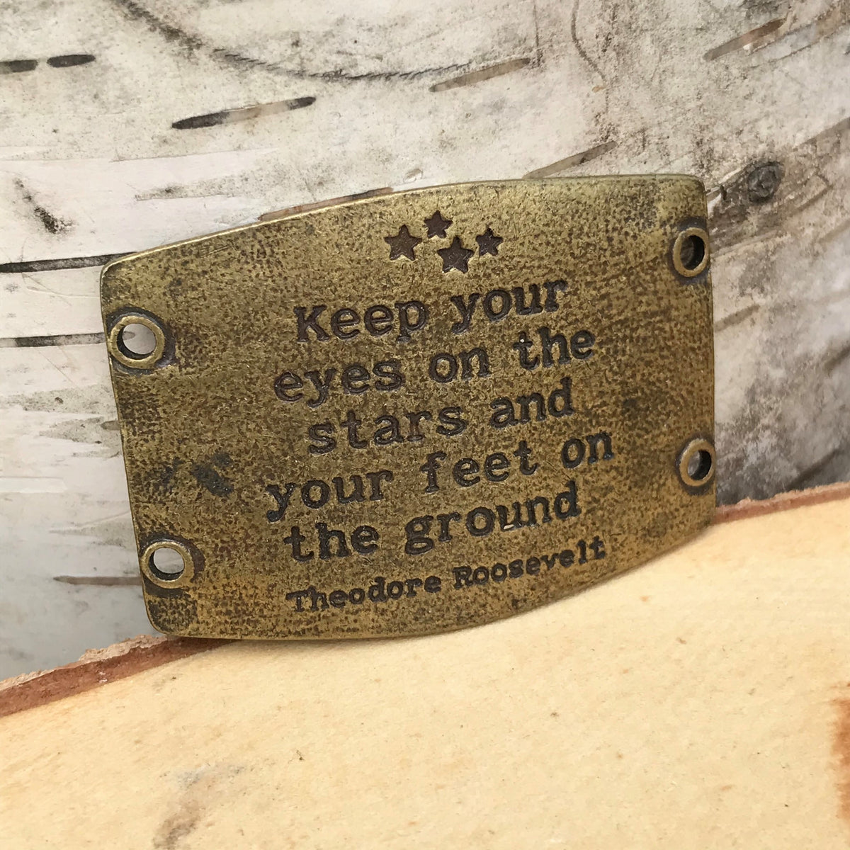 Antique brass finish Lenny & Eva bracelet sentiment that reads, "Keep your eyes on the stars and your feet on the ground." Quote by Theodore Roosevelt. Design of stars above the quote.