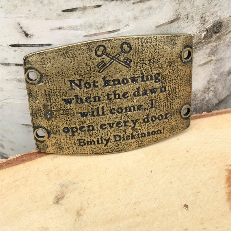 Antique brass finish Lenny & Eva bracelet sentiment that reads, "Not knowing when the dawn will come, I open every door." Quote by Emily Dickinson. A set of keys is etched above the quote.