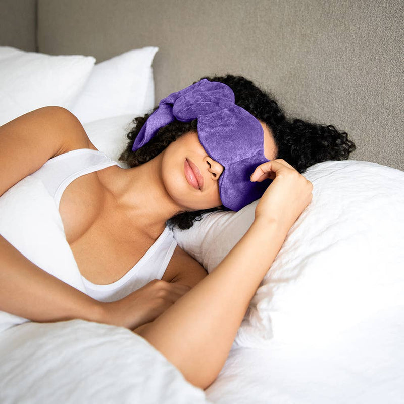 Woman laying on her side in bed with a purple weighted eye mask over her eyes